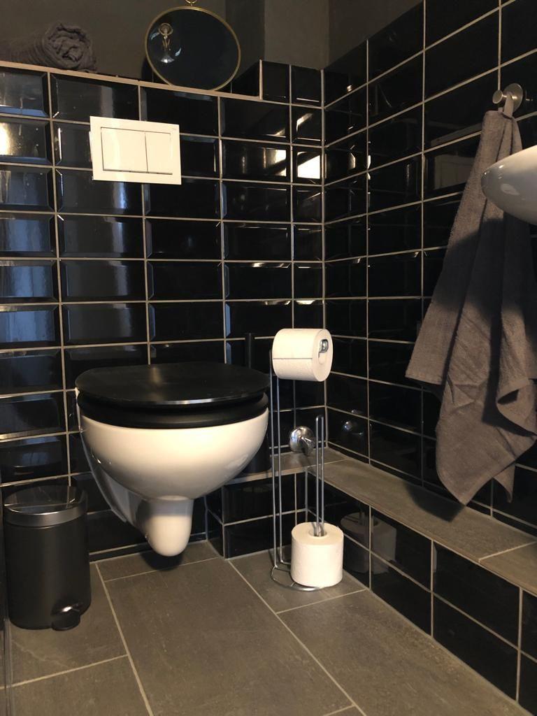 Toilette mit schwarzen Fliesen und grauem Boden. Toilettenpapier und Handtuch sind vorhanden.