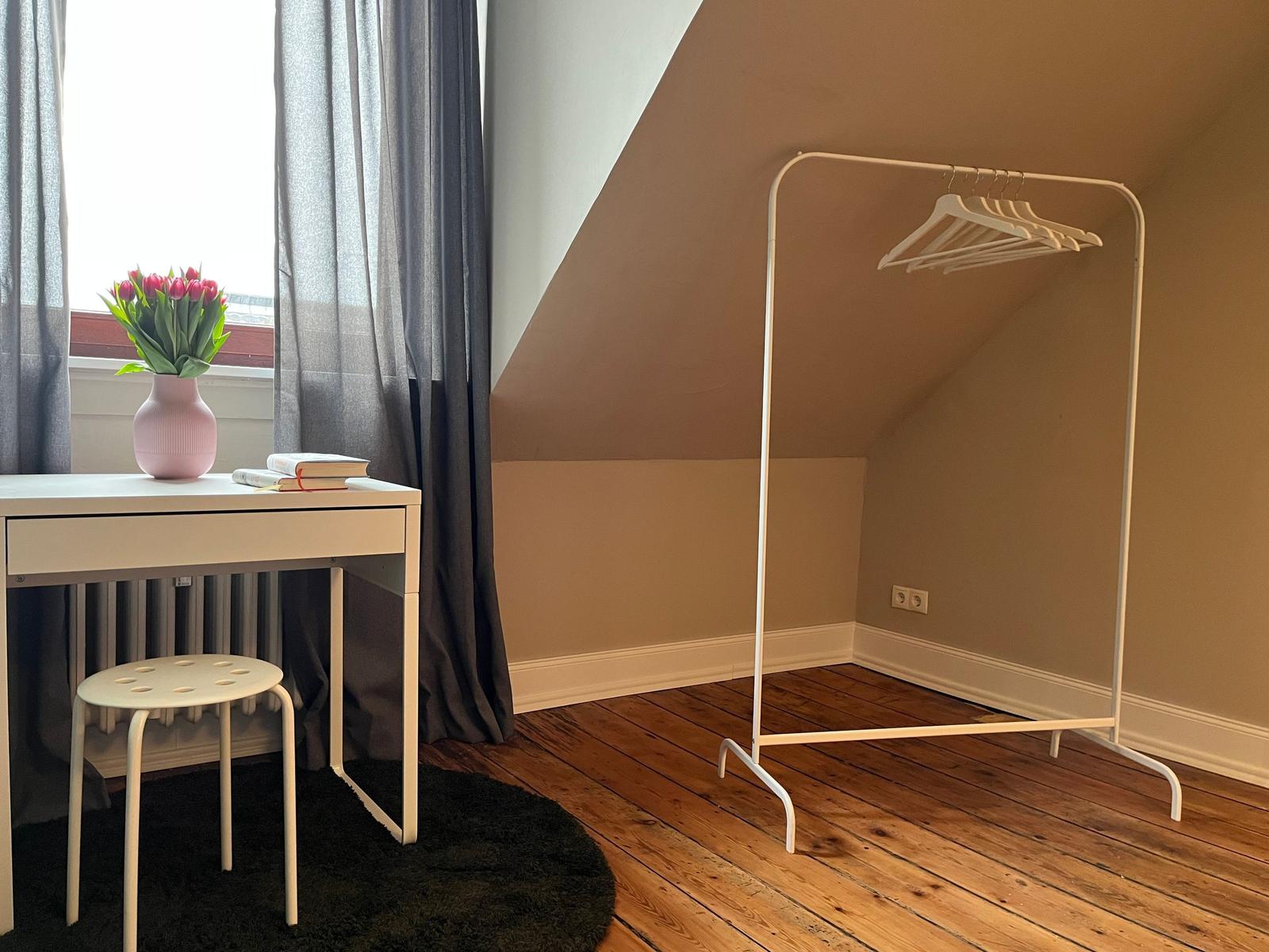 Schlafzimmer mit Schreibtisch, Stuhl und Kleiderständer unter Dachschrägen.