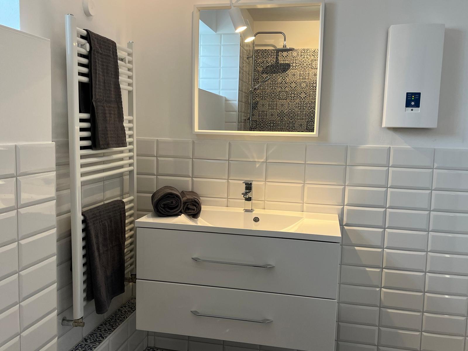 Badezimmer mit Waschbecken, Spiegel und Dusche. Weiße Fliesen und Handtuchhalter.