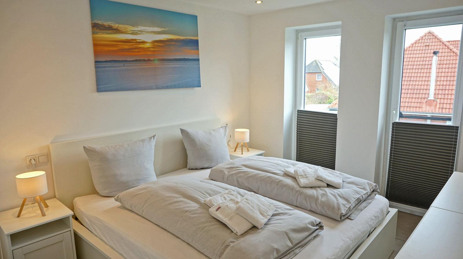 Schlafzimmer mit Doppelbett, zwei Nachttischen und Fenstern mit Blick auf Dächer.