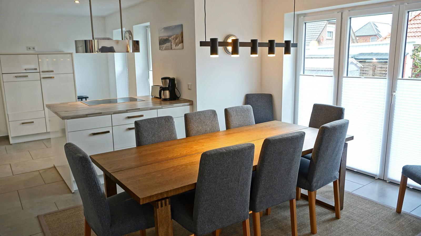 Essbereich mit Holztisch und grauen Stühlen. Küche mit Arbeitsplatte und Fenster.
