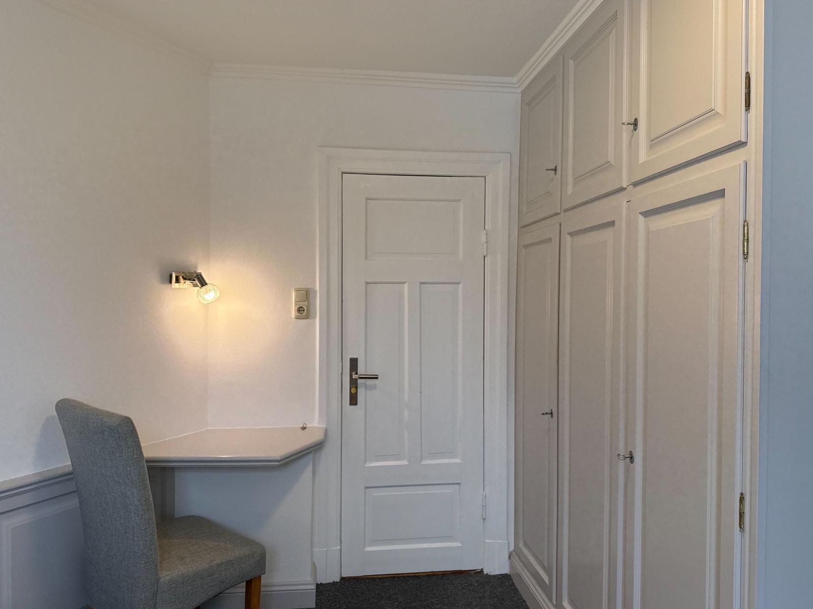 Room with desk, chair, and wardrobe. Door and wall light visible.