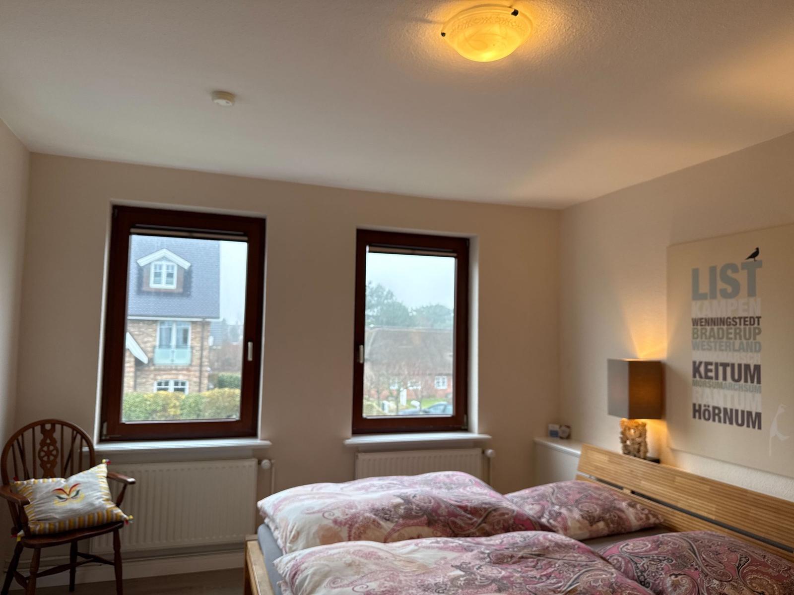 Schlafzimmer mit Doppelbett, zwei Fenstern und Wandbild.