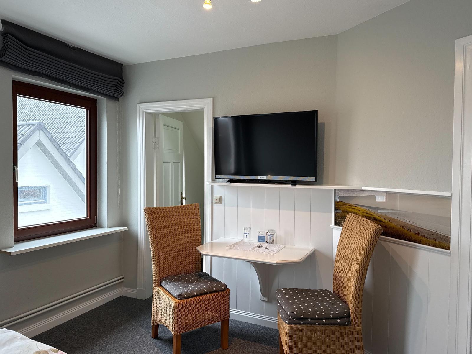 Room with TV, desk and two chairs. Window with view of roof.