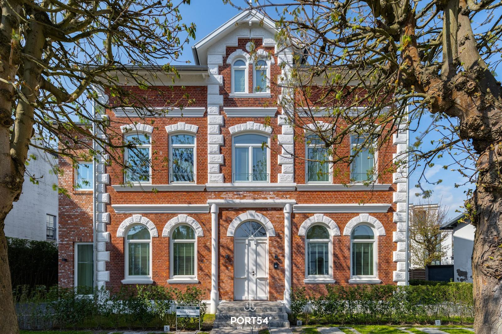 Rotes Backsteinhaus mit weißen Fensterrahmen und Eingangstür. Umgeben von Bäumen und Grünflächen.