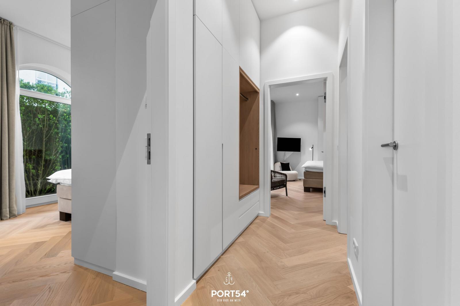 Hölzerner Boden, weiße Schränke und Blick in Schlafzimmer mit Bett und Fernseher.