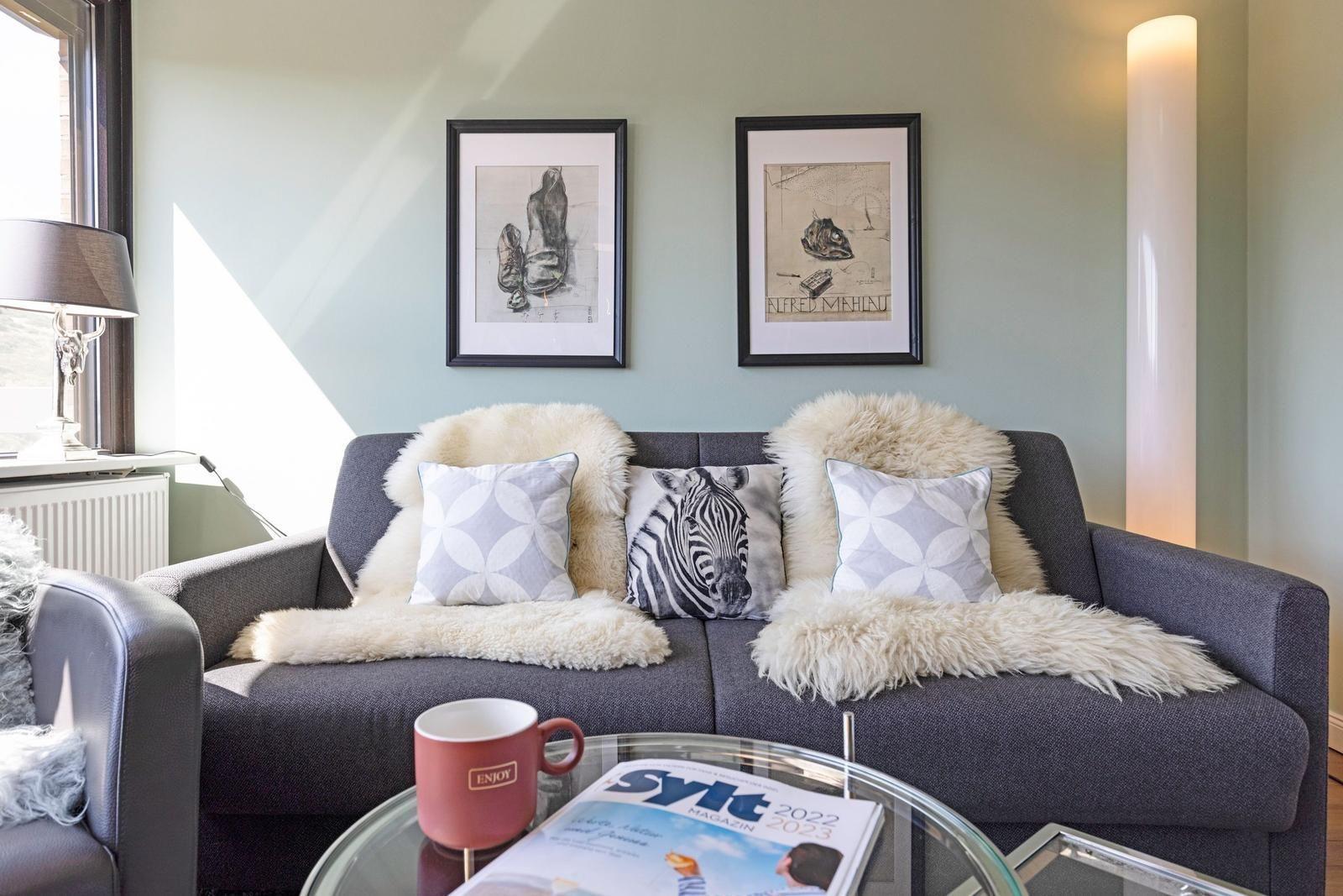 Ein grauer Sofa mit Felldecken und Kissen. Auf dem Tisch steht eine Tasse und ein Magazin.