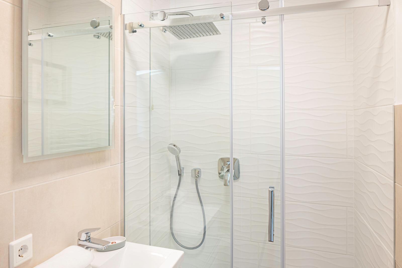 Dusche mit Glaswand, Duschbrause und Handdusche. Spiegel und Waschbecken daneben.