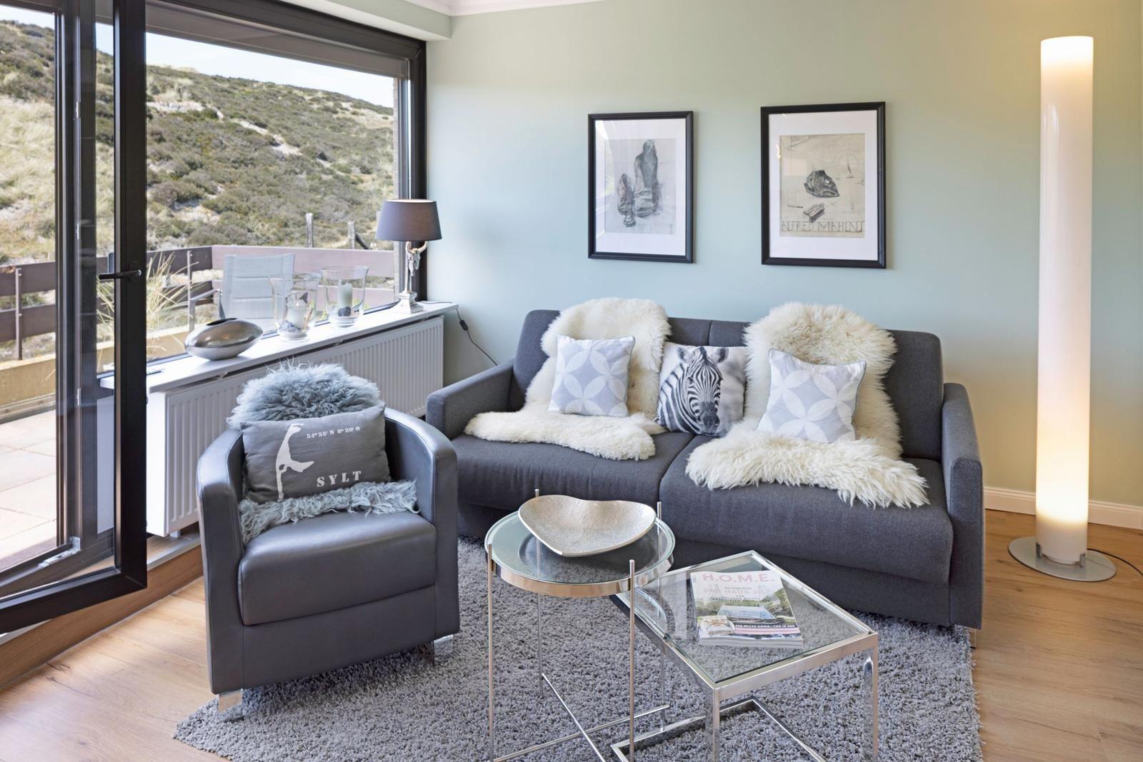 Ein Wohnzimmer mit grauem Sofa, Sessel und Glas-Tischen. Blick auf eine Terrasse mit Berglandschaft.