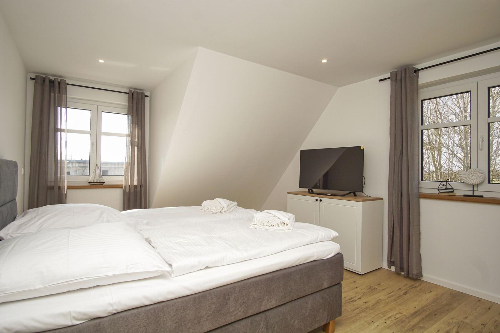 Schlafzimmer mit Doppelbett, Fernseher und Fenstern mit Vorhängen.