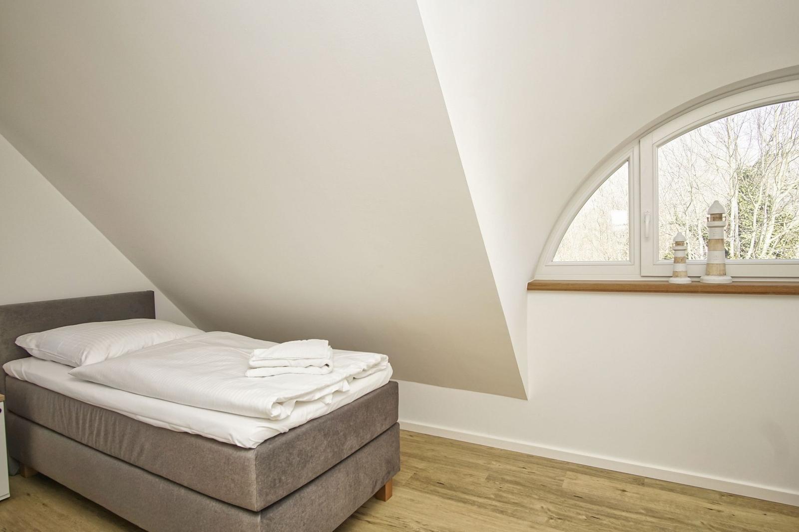 Schlafzimmer mit Dachschräge, Bett und Bogenfenster mit Blick auf Bäume.