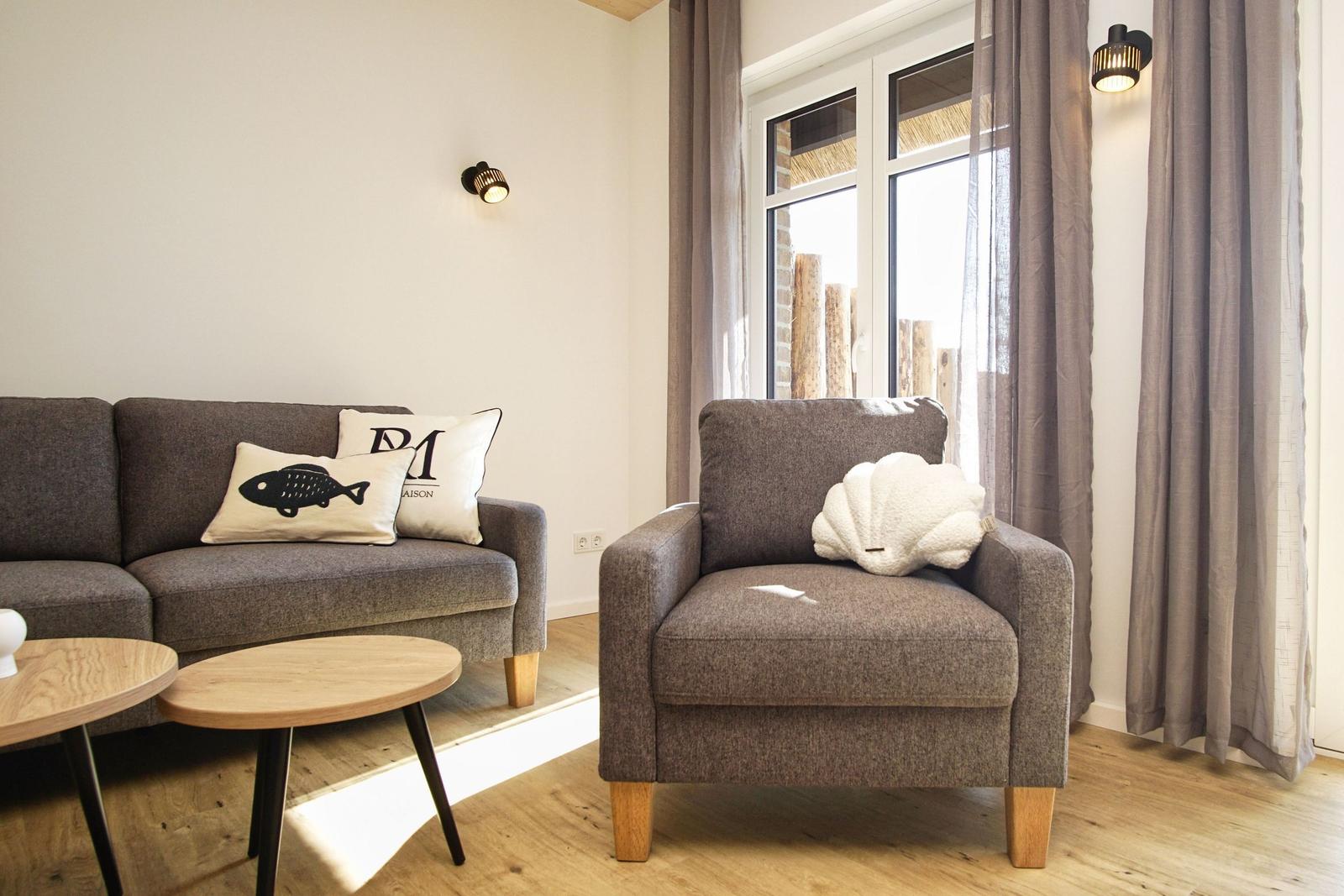 Ein Wohnzimmer mit grauem Sofa und Sessel, Holztisch und Fenster mit Vorhängen.