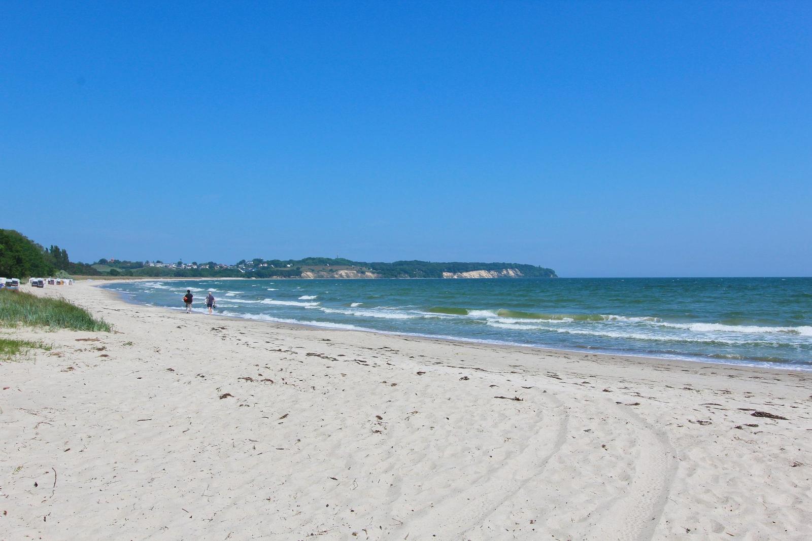 Strand mit weißen Sand, Wellen und klarem Himmel. Zwei Personen spazieren entlang des Wassers.