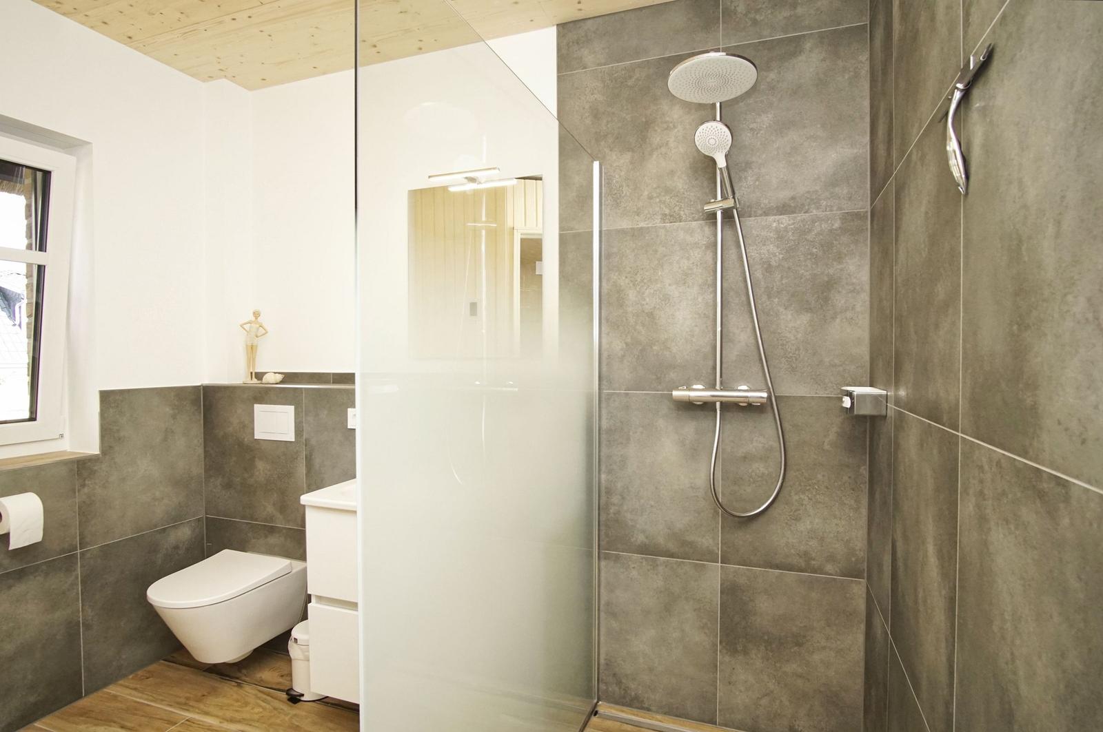 Badezimmer mit Dusche, WC und Fenster. Graue Fliesen und Holzdecke.
