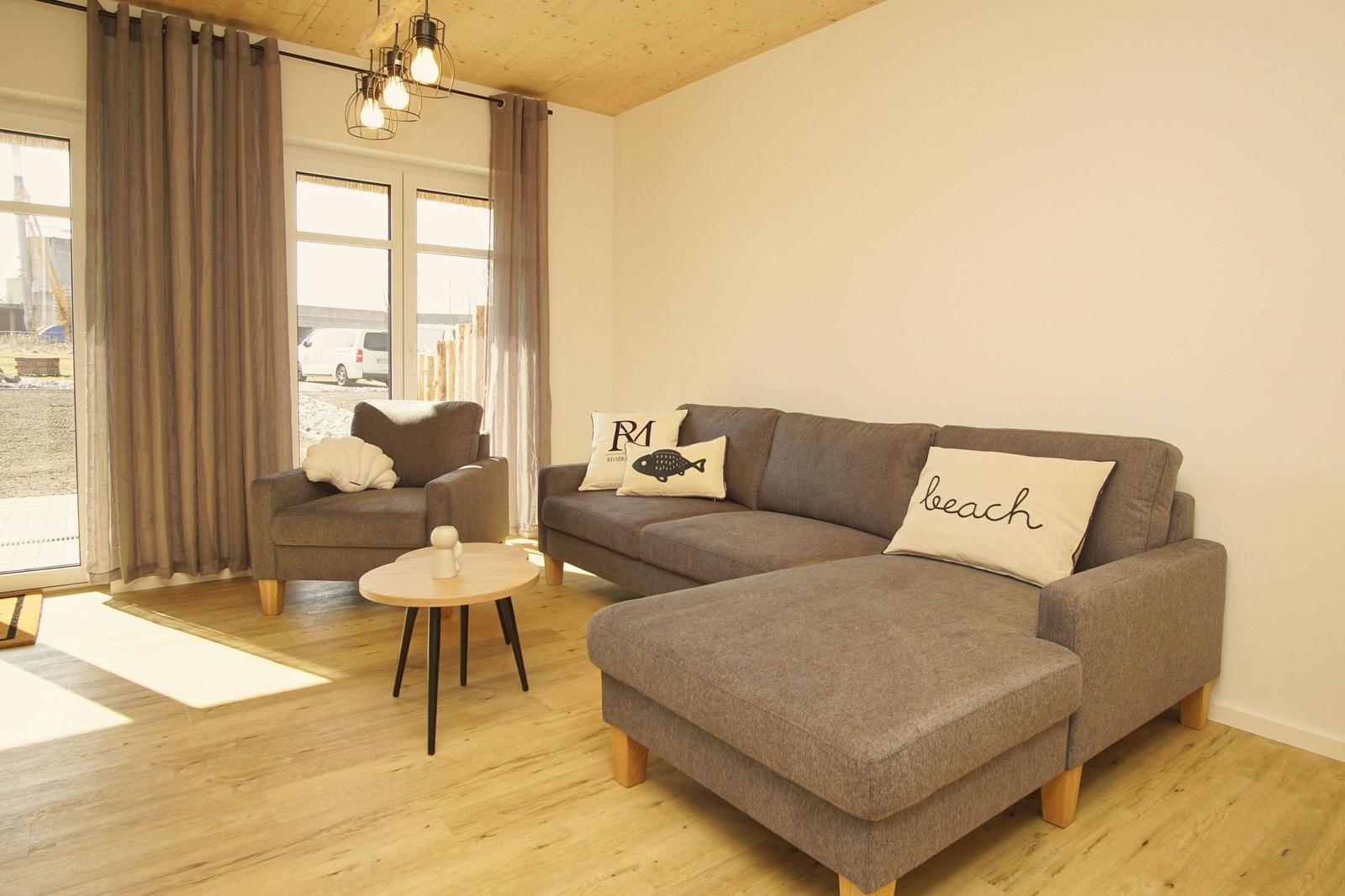 Wohnzimmer mit grauem Sofa, kleinem Tisch und Fenstern mit Vorhängen.