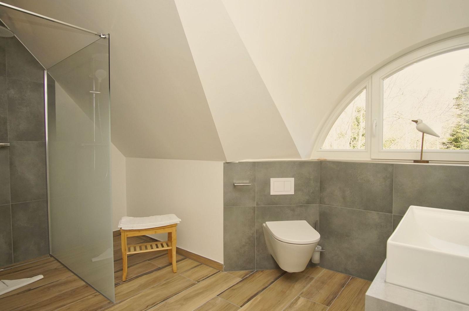 Badezimmer mit Dusche, Toilette und Waschbecken unter Dachschräge mit Fenster.