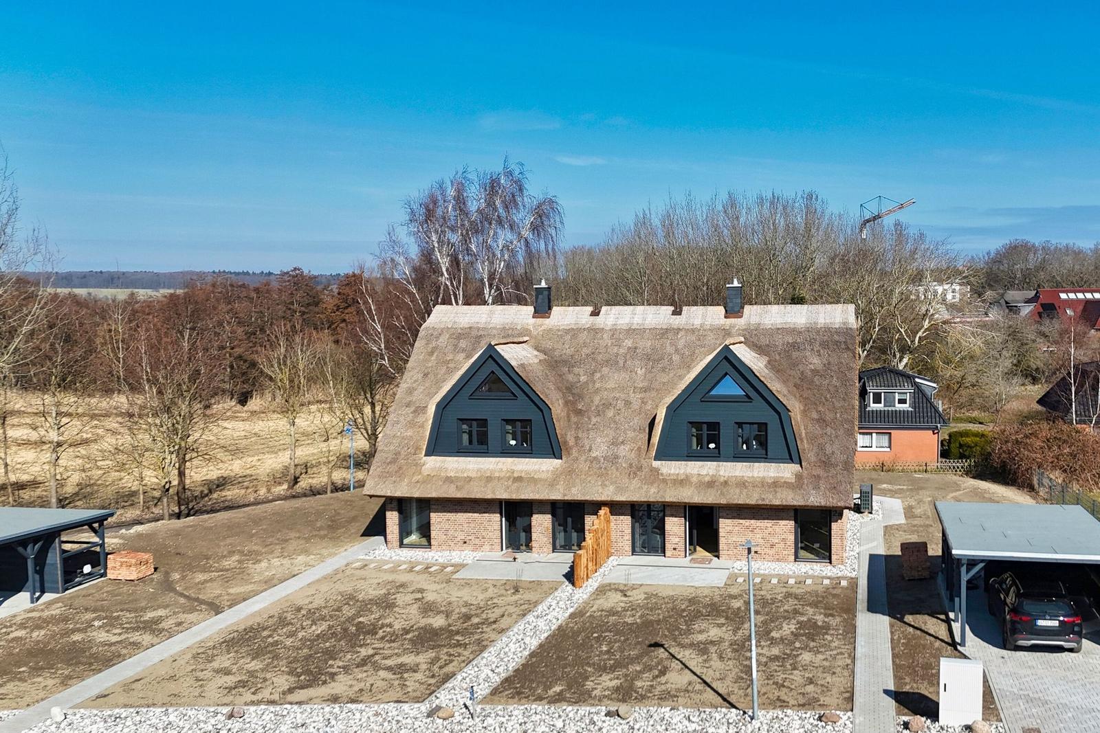 Modernes Haus mit Strohdach, Terrasse und Parkplatz. Umgeben von Wald und Feldern.