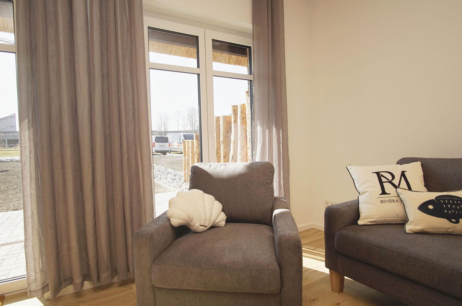 Ein Wohnzimmer mit zwei Sitzmöbeln und großen Fenstern. Auf den Möbeln liegen Kissen.