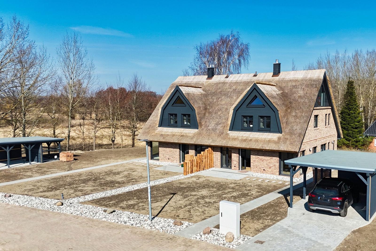Modernes Haus mit Strohdach und Carport. Freier Platz und Bäume im Hintergrund.