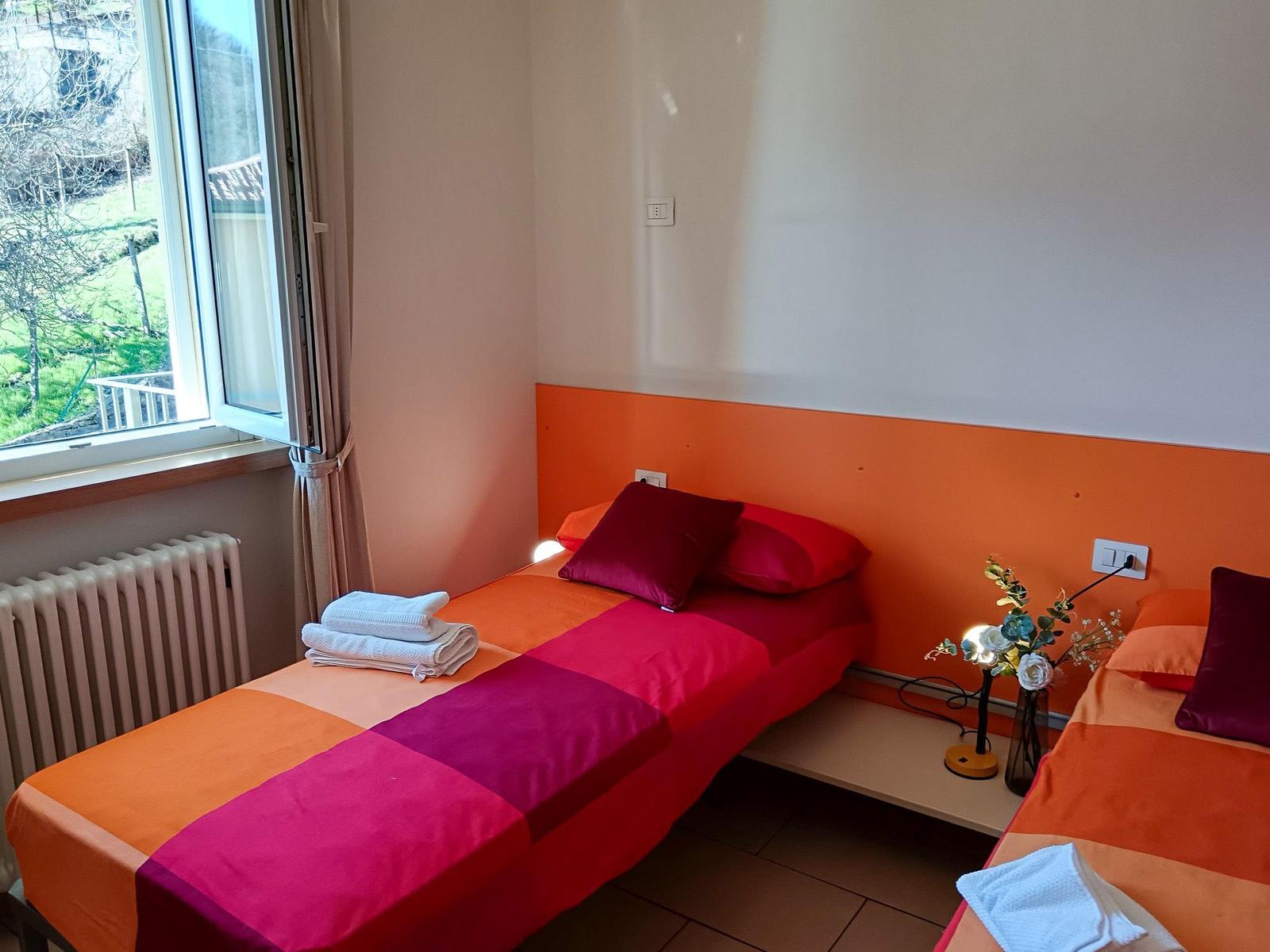 Zwei Betten mit bunten Bettwäschen, Fenster mit Blick auf Grünfläche und Heizkörper.