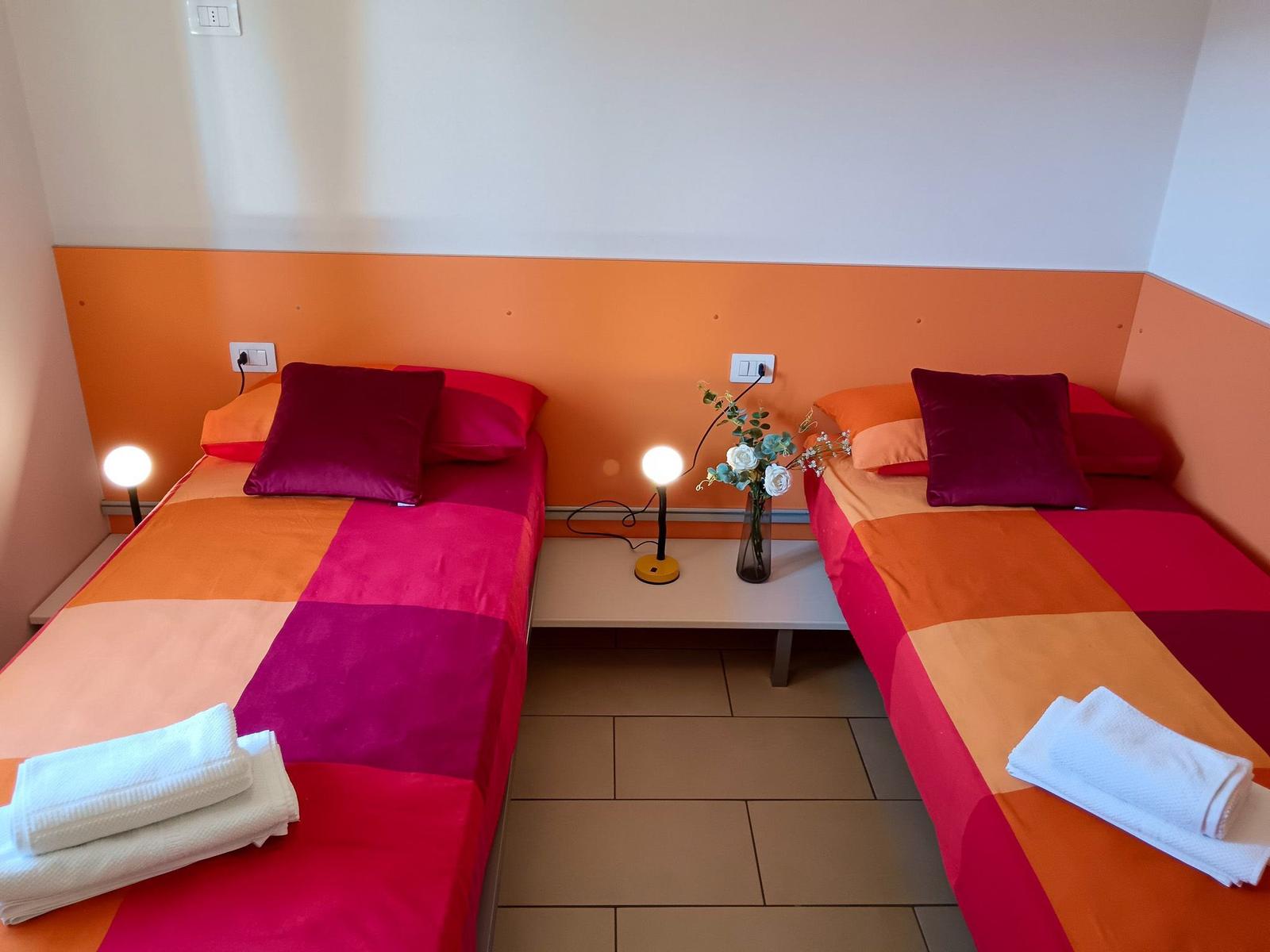 Zwei Betten mit bunten Bettwäschen und einer Tischlampe in einem Raum mit orangefarbener Wand.