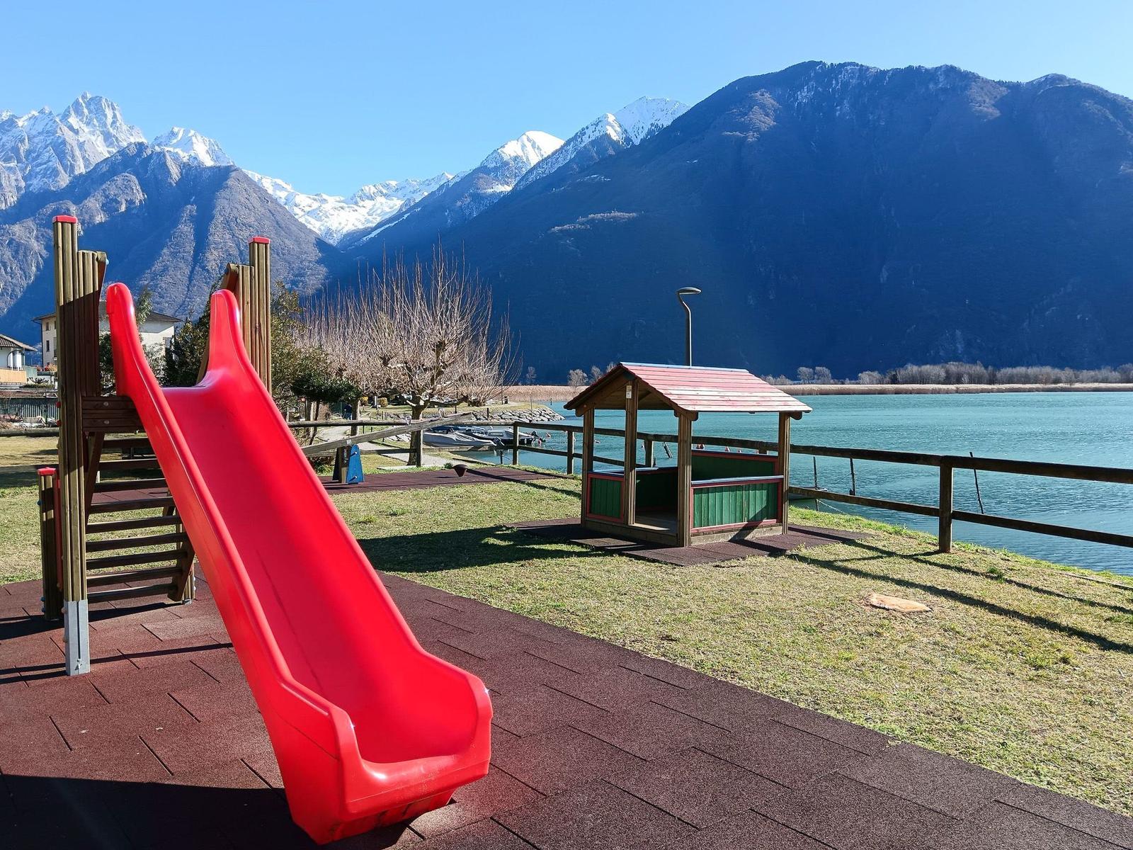 Roter Rutschbahn und Spielhütte am See mit Bergen im Hintergrund.