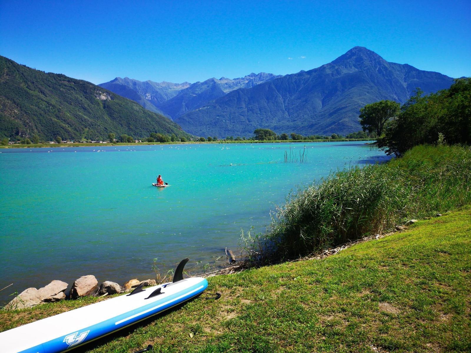 Ein Stand-Up-Paddleboard liegt am Ufer eines türkisfarbenen Sees mit Bergen im Hintergrund.
