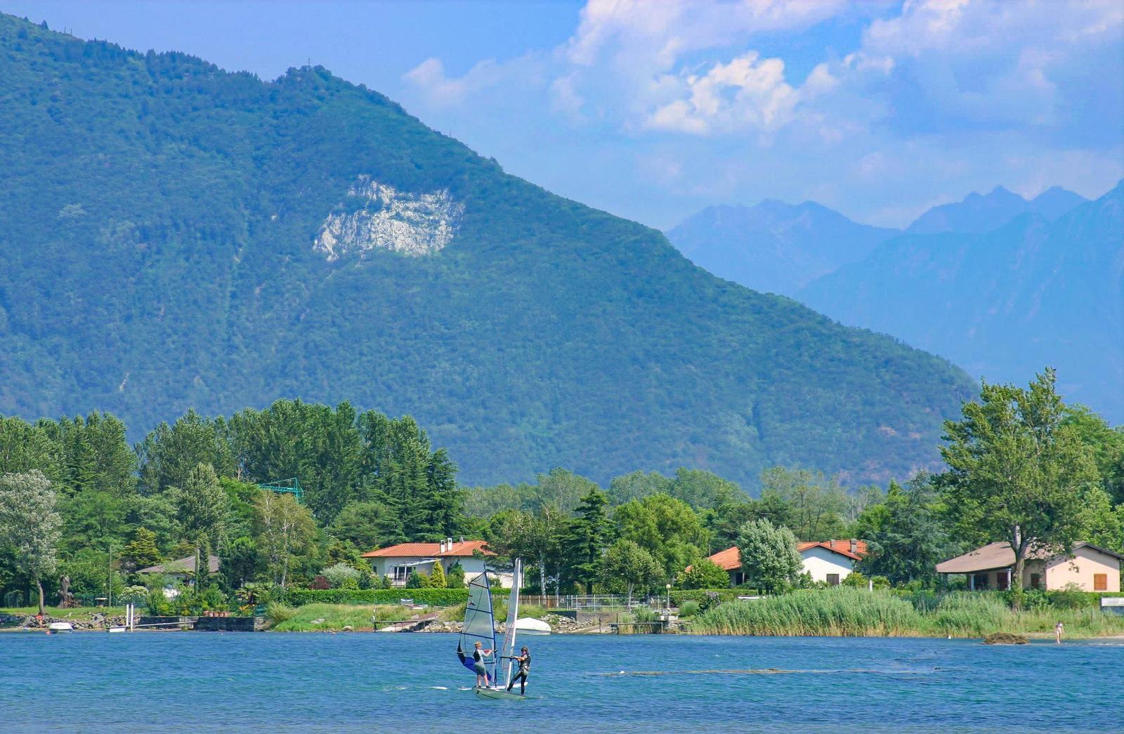 Zwei Personen segeln auf einem See vor grünen Bergen und Häusern am Ufer.