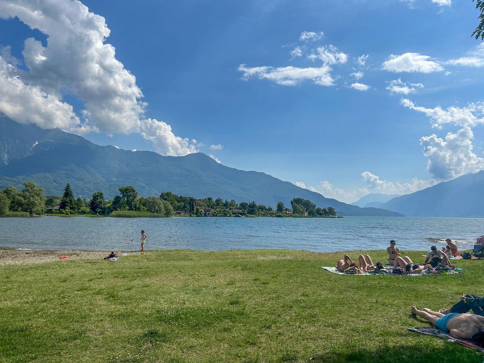 Grüner Strandbereich am See mit Menschen, die sich entspannen und die Berge im Hintergrund.
