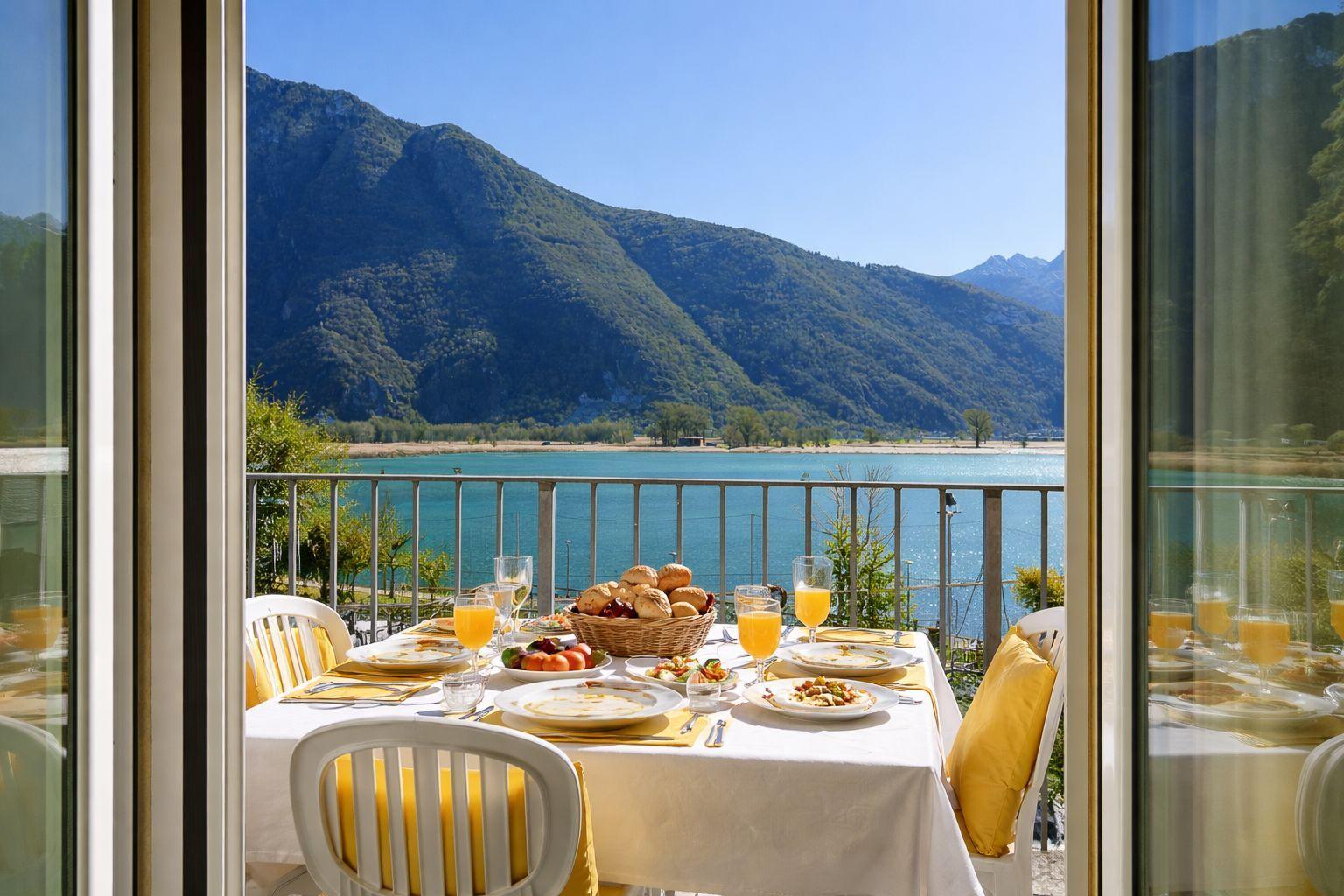 Balkon mit Tisch, Stühlen und Frühstück. Blick auf See und Berge.