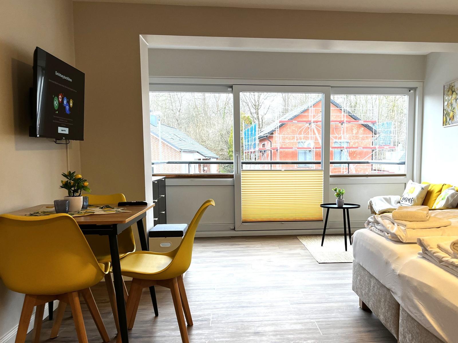 Wohnraum mit Essbereich, TV und Balkon mit Blick auf Gebäude.
