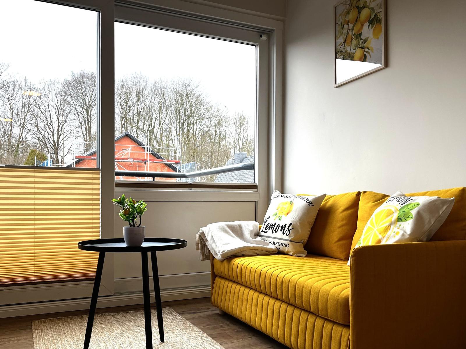 Gelber Sofa mit Kissen, kleiner Tisch und Fensterblick auf Bäume.