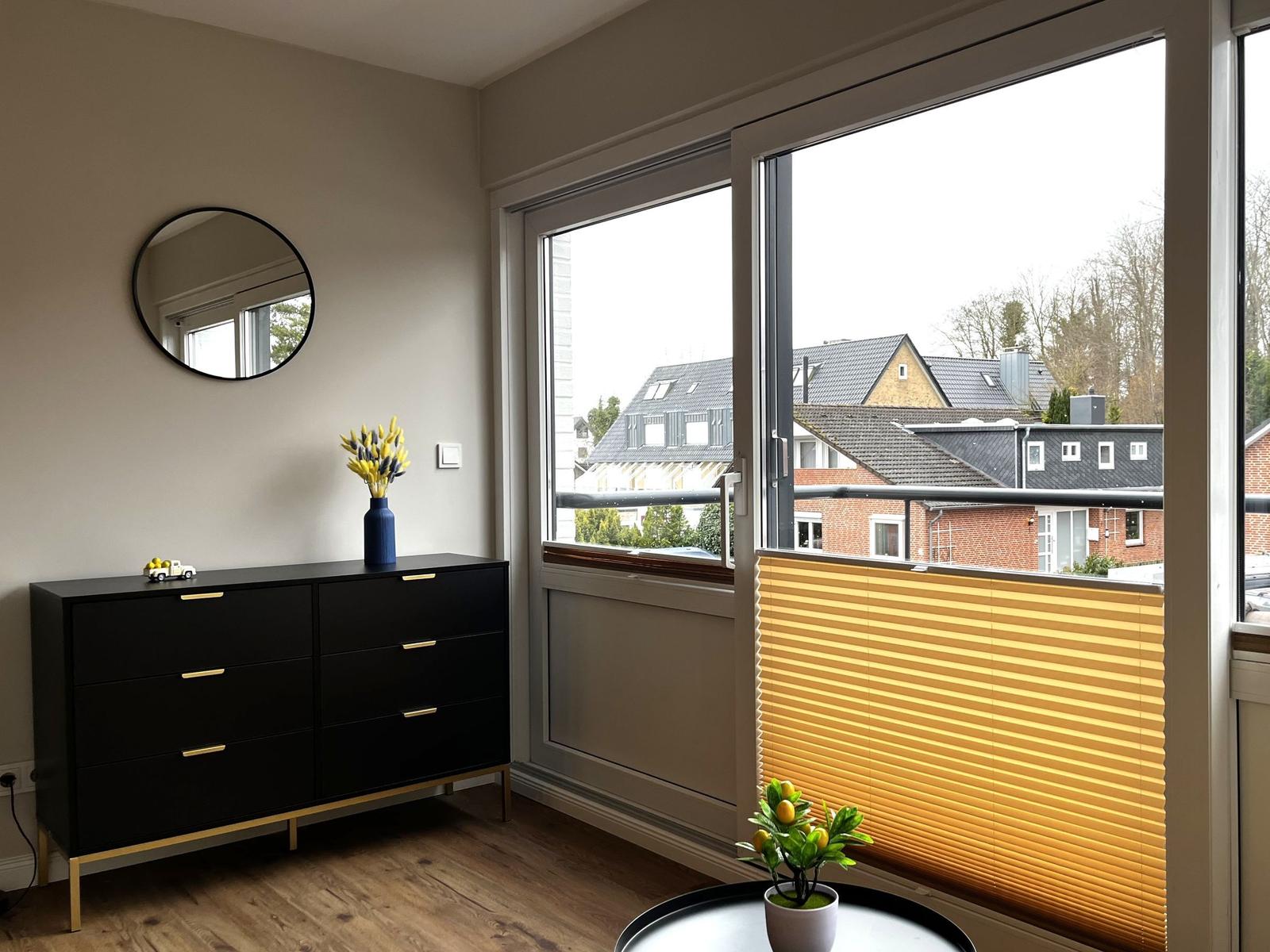Schwarzer Schrank mit goldfarbenen Griffen und runder Spiegel an der Wand. Fenster mit Jalousie und Blick auf Dächer.