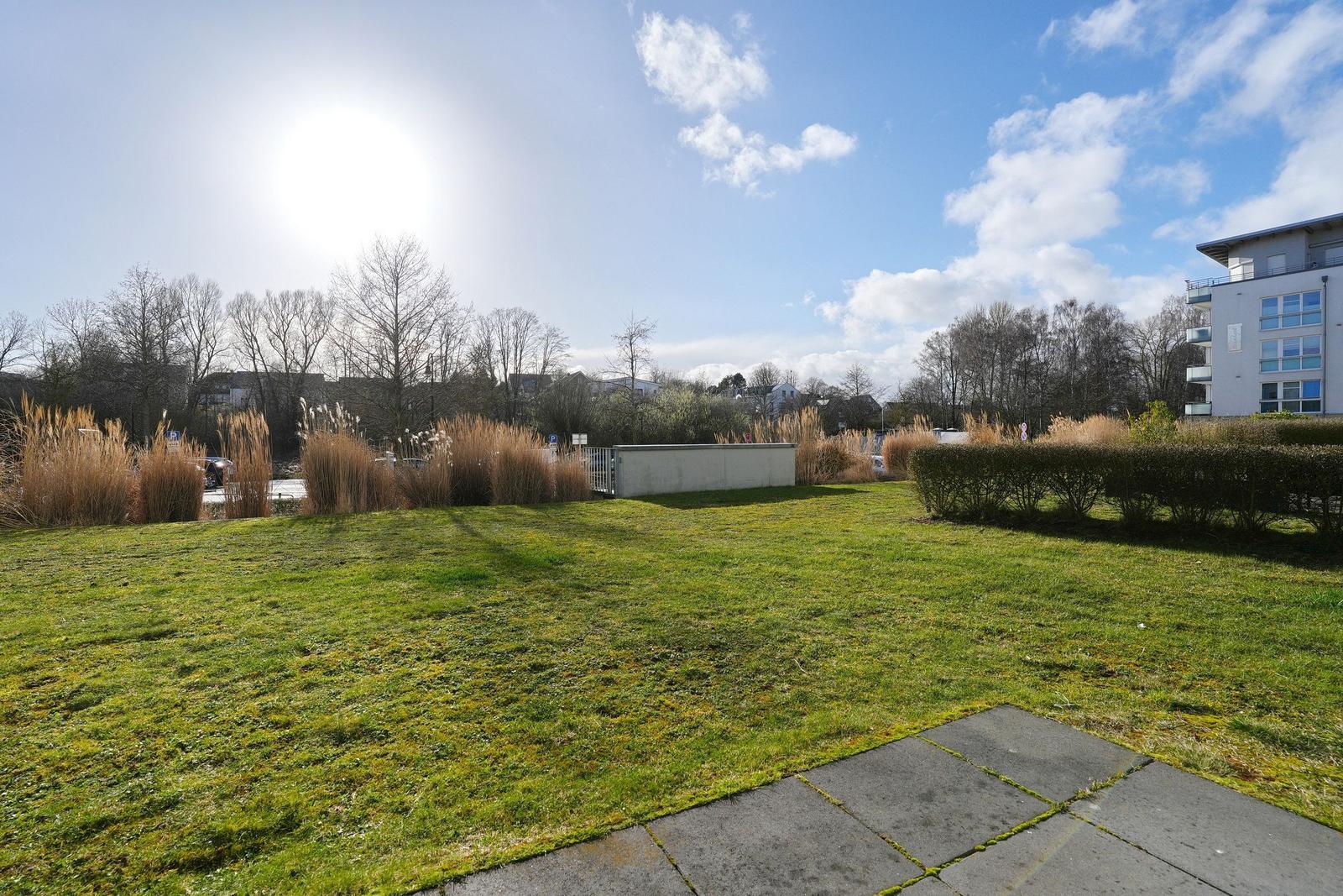 Grüner Rasenbereich mit Weg, Hecken und Bäumen unter blauem Himmel.