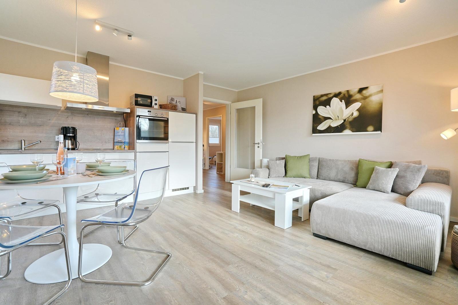 Wohn- und Essbereich mit Küchenzeile und Sofa. Holzboden und helle Wände.