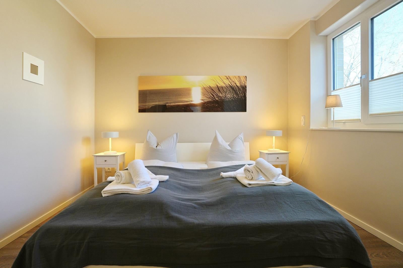 Schlafzimmer mit Bett, zwei Nachttischen und Wandbild. Fenster mit Jalousie.