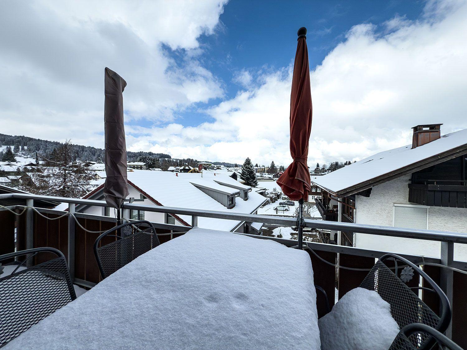 Schneebedeckter Balkon mit Tisch und Stühlen, Blick auf verschneite Dächer und Berge.