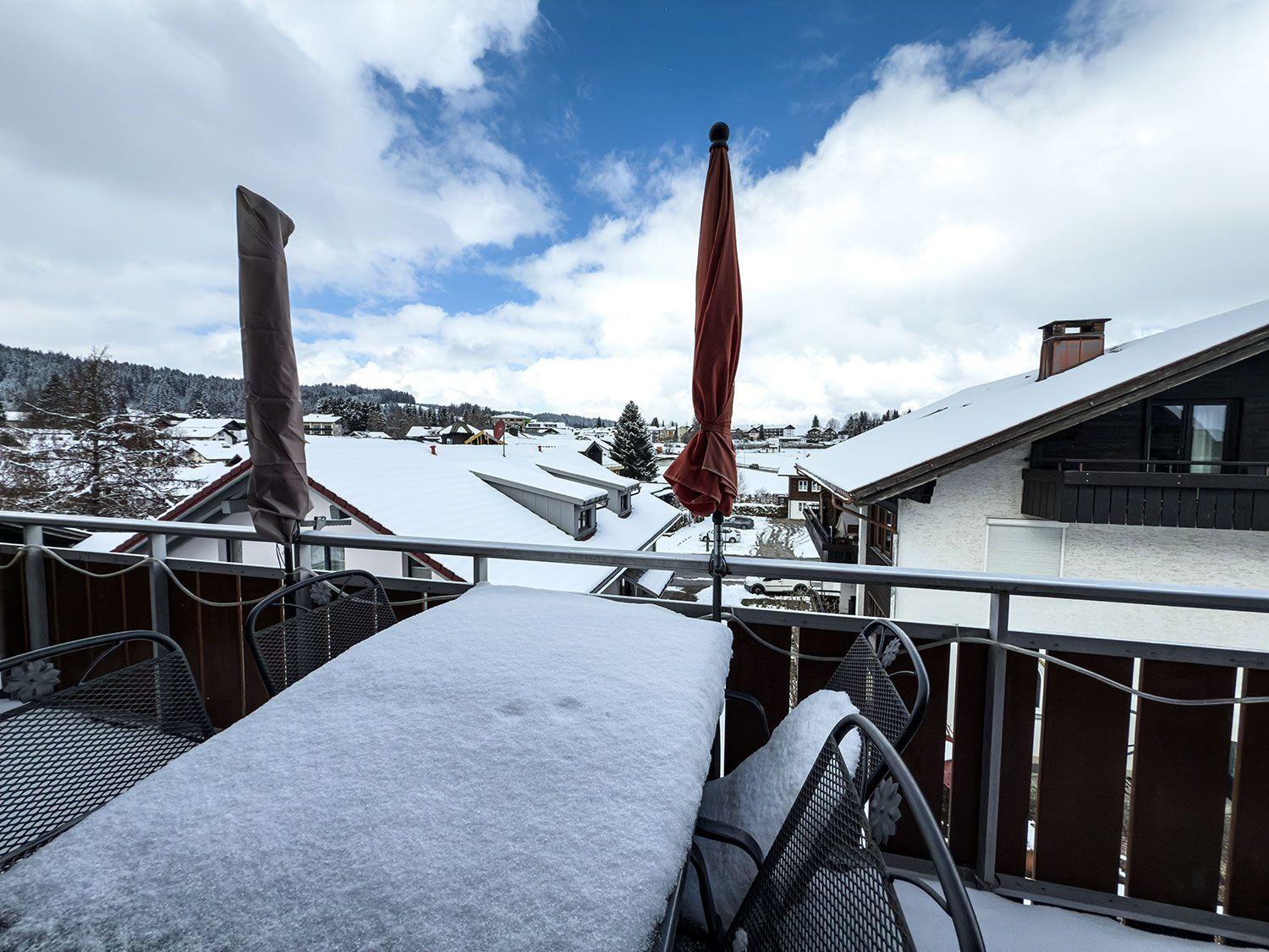 Schneebedeckter Balkon mit Tisch und Stühlen, Blick auf schneebedeckte Dächer und Berge.