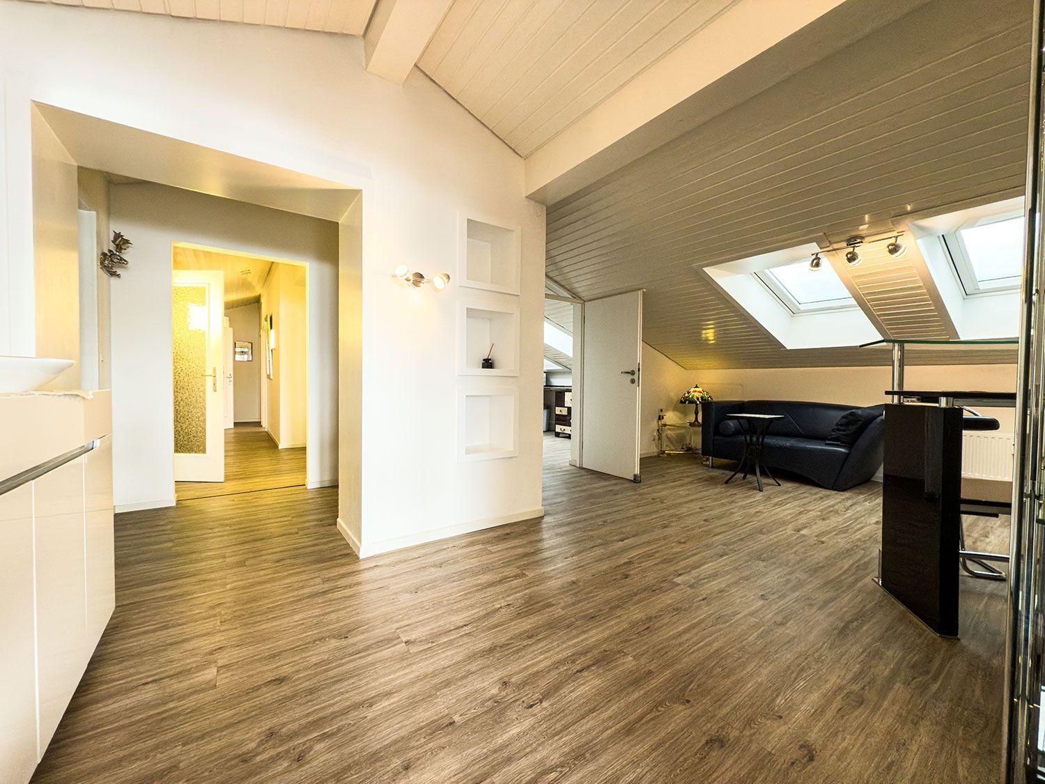 Wohnung mit Holzboden, Dachschrägen und Fenstern. Ein dunkler Sofa und eine Küche sind sichtbar.