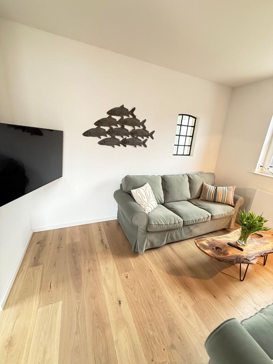 Living room with sofa, TV, and wooden table. Wall decoration and windows visible.