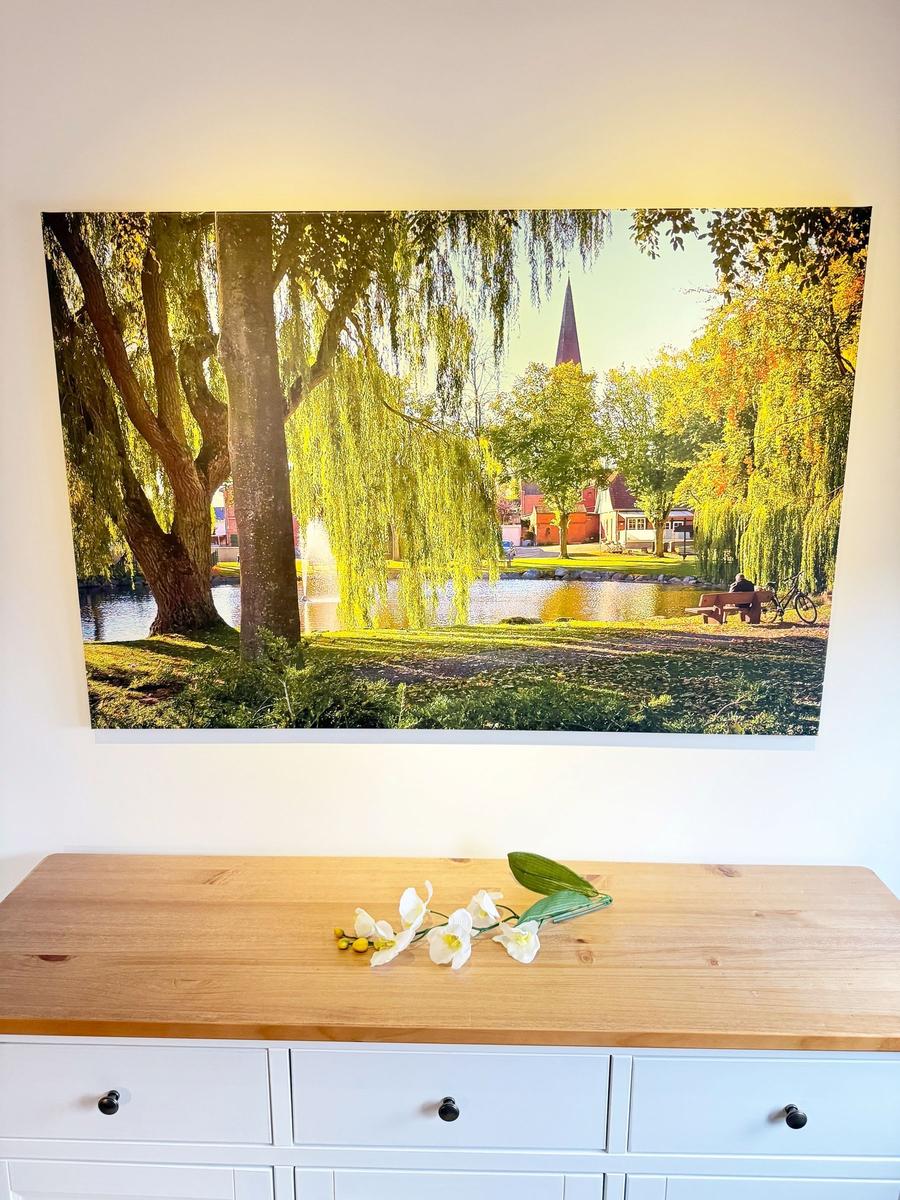 Wall picture shows a lake with trees and church tower. White flowers lie on the table.