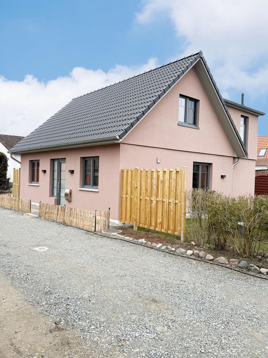 A pink house with gray roof and wooden fence. Gravel yard with shrubs.
