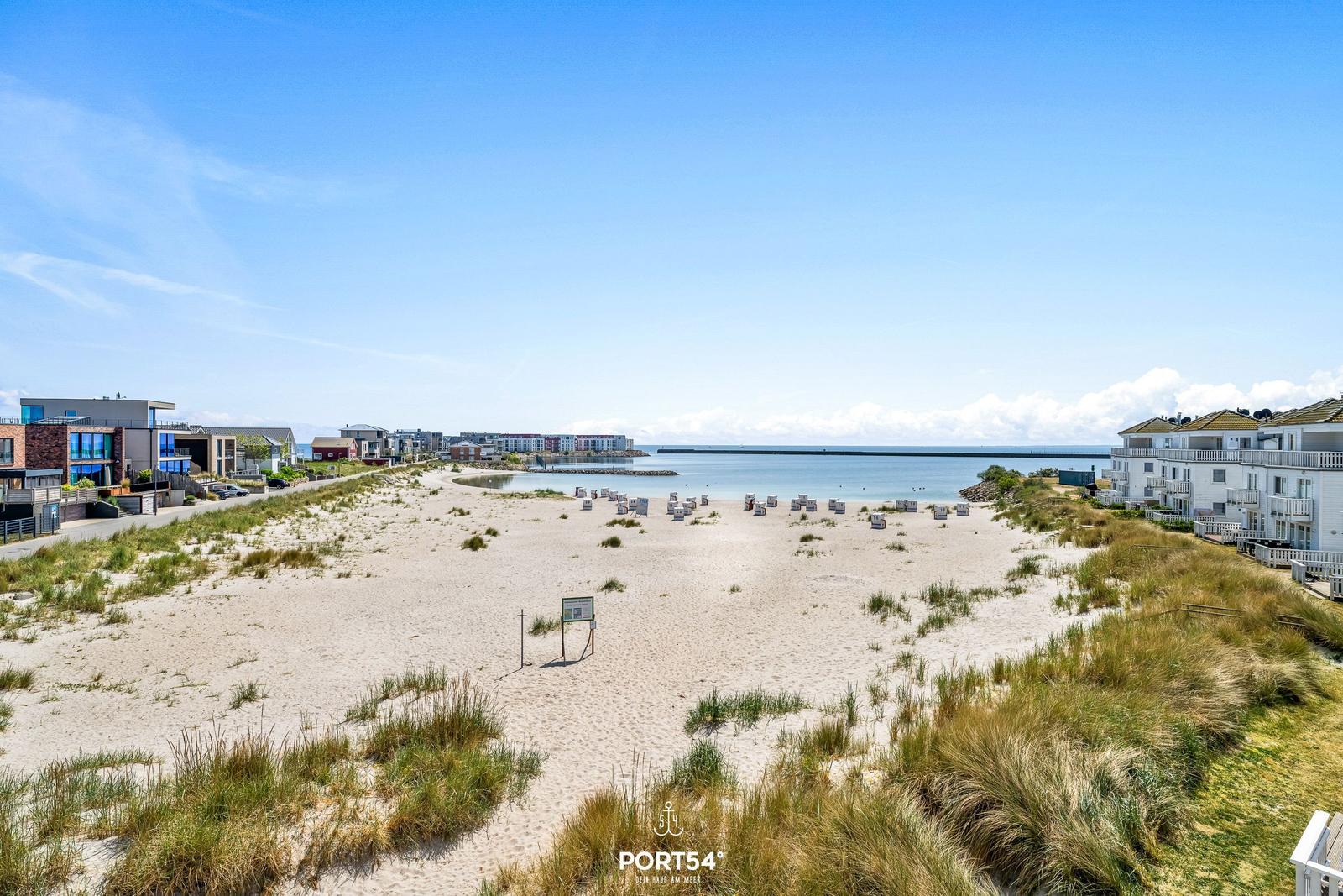 Strand mit Sanddünen, Häusern und Blick aufs Meer unter blauem Himmel.