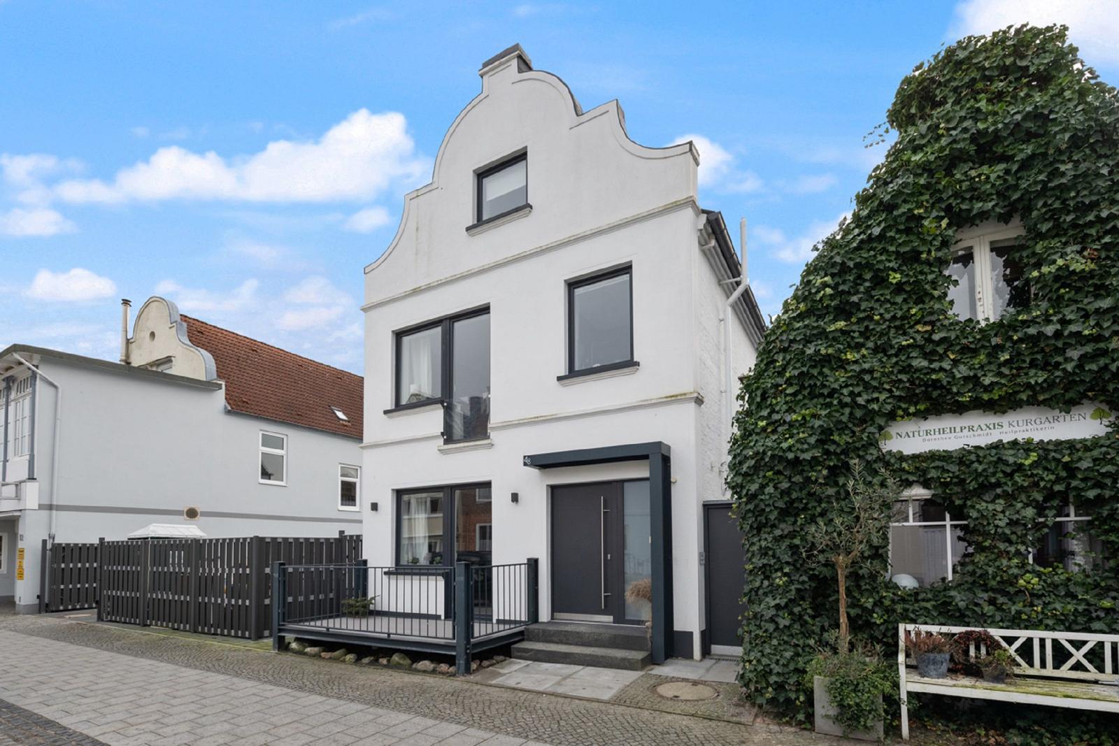 Weiße Hausfassade mit dunklen Fenstern und Eingang. Rechts wächst Efeu an einem Gebäude.