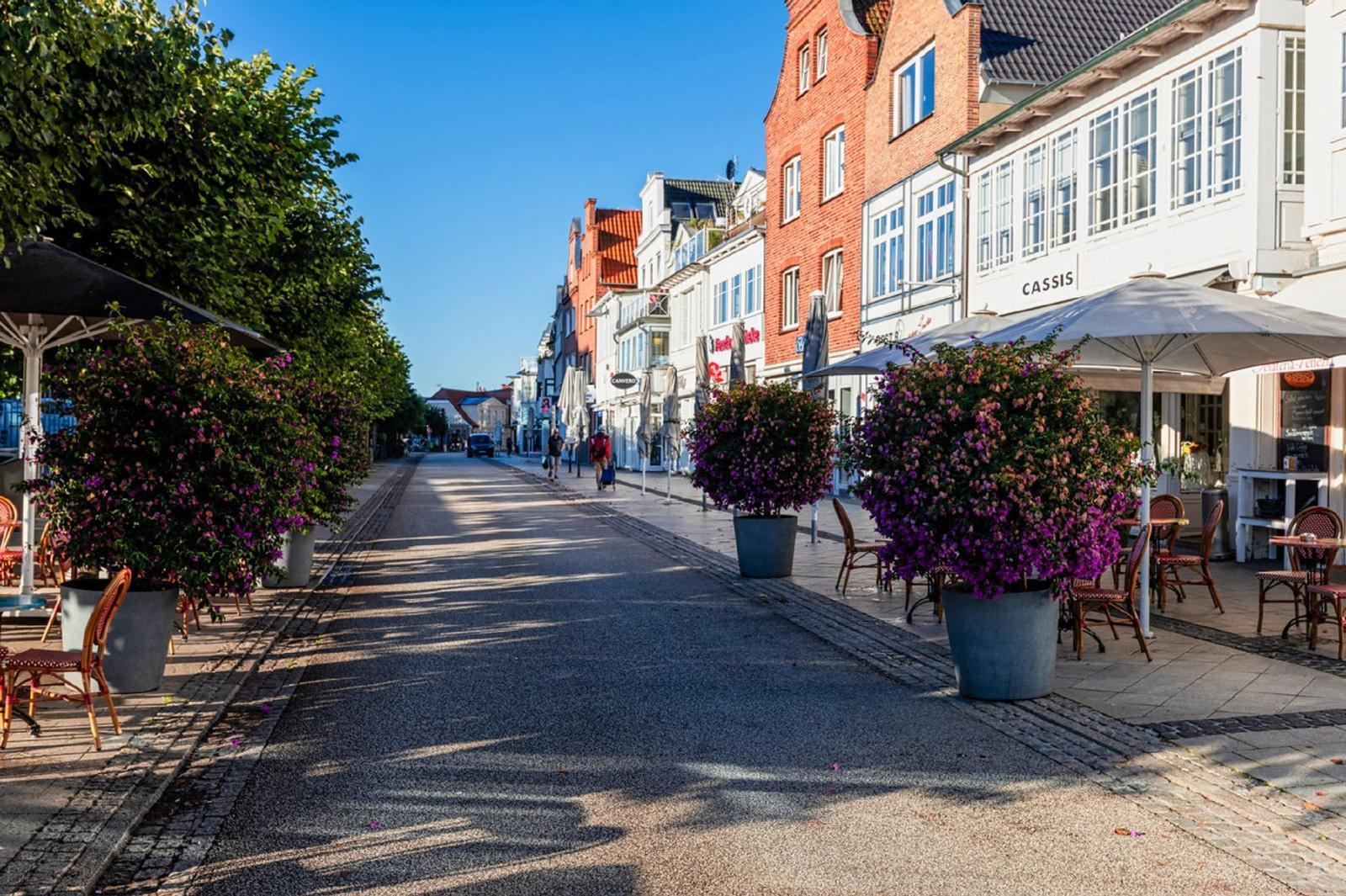 Straße mit Blumenkübeln und Caféterrassen unter klarem Himmel.