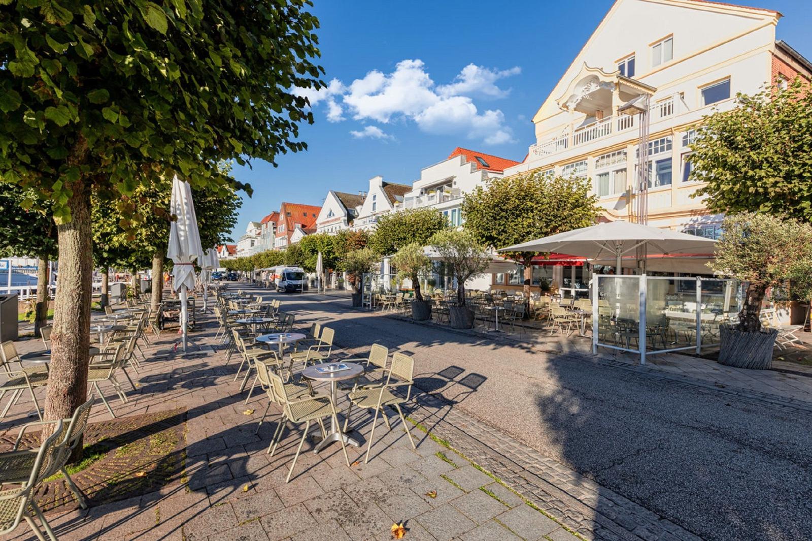 Außenbereich mit Tischen, Stühlen und Sonnenschirmen entlang einer Straße mit Bäumen und Gebäuden.