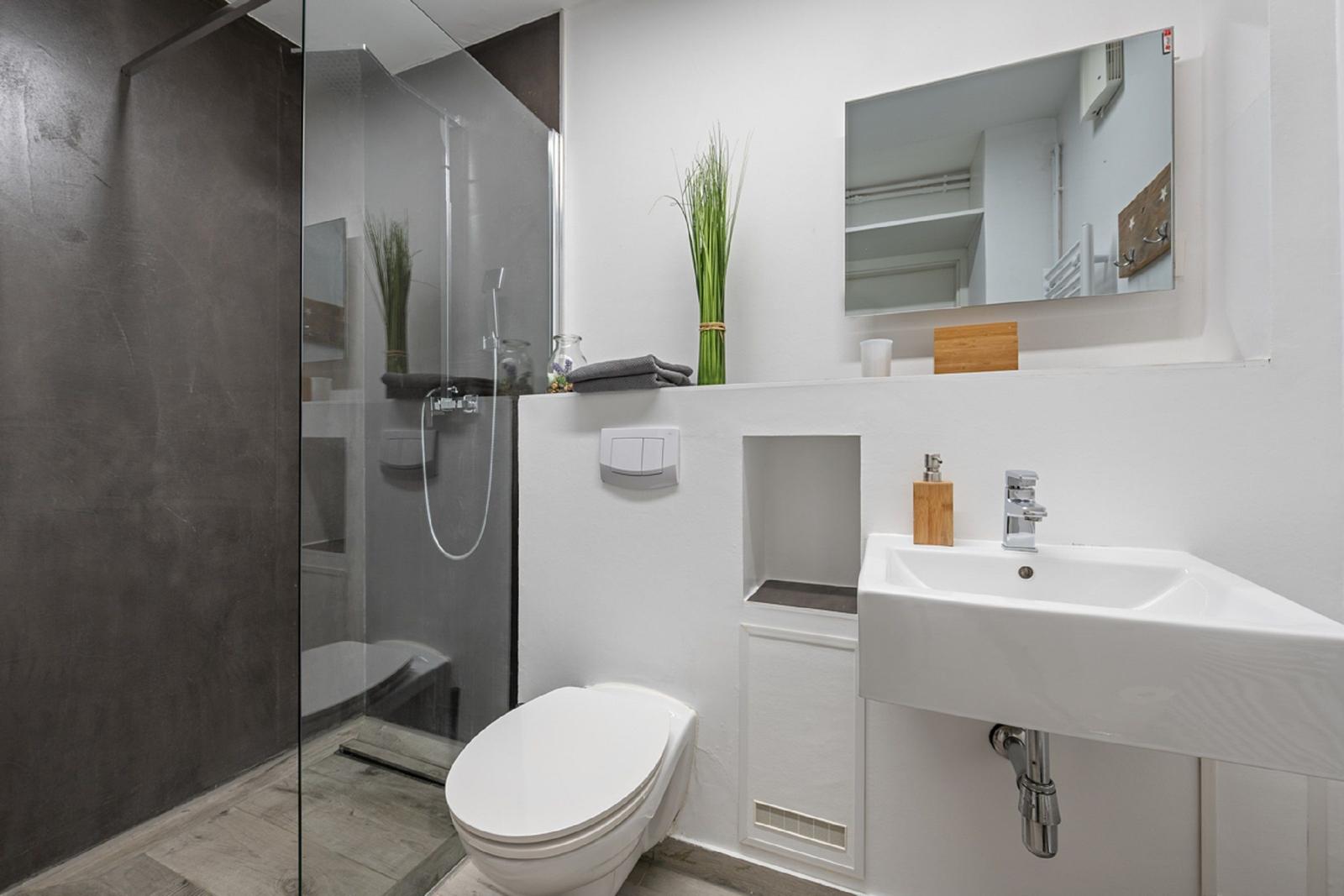 Badezimmer mit Dusche, Waschbecken und Toilette. Weiße Wände und graue Fliesen.