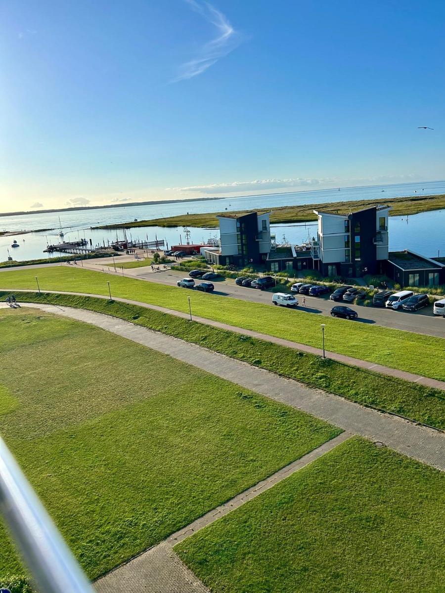 Hafenansicht mit modernen Gebäuden und Parkplatz. Grünflächen und Wasser im Hintergrund.