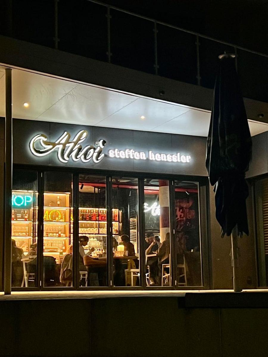 Außenansicht von Ahoi steffen henssler mit beleuchteter Fassade und Sitzgelegenheiten im Inneren.
