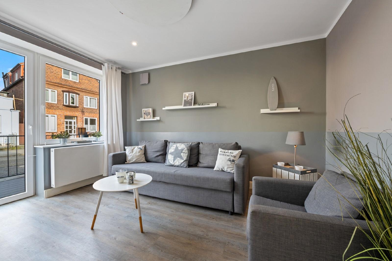 Wohnzimmer mit grauem Sofa, weißen Regalen und Blick auf Gebäude.